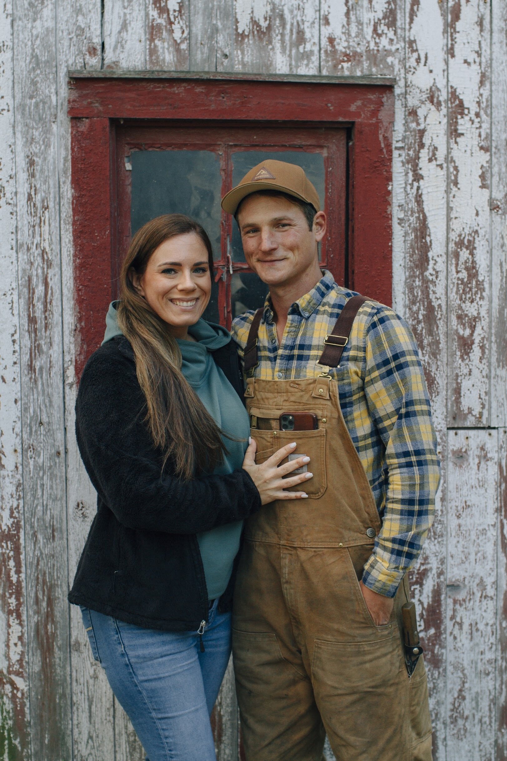 Jacobson+Family+Farms+Illinois+-+-MG-1061-2 Young couple posing together in front of a rustic barn, showcasing a warm and friendly atmosphere. The woman wears a black jacket and blue jeans, while the man is dressed in a plaid shirt and brown overalls, exuding a casual, farm-inspired style. The weathered wooden background adds to the charm of the rural setting.