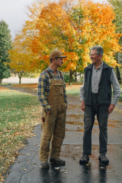 Two men engaged in conversation on a rainy autumn day, surrounded by colorful fall foliage. The younger man wears a plaid shirt and overalls, while the older man is dressed in a vest and trousers. The scene captures the essence of rural life and intergenerational bonding.