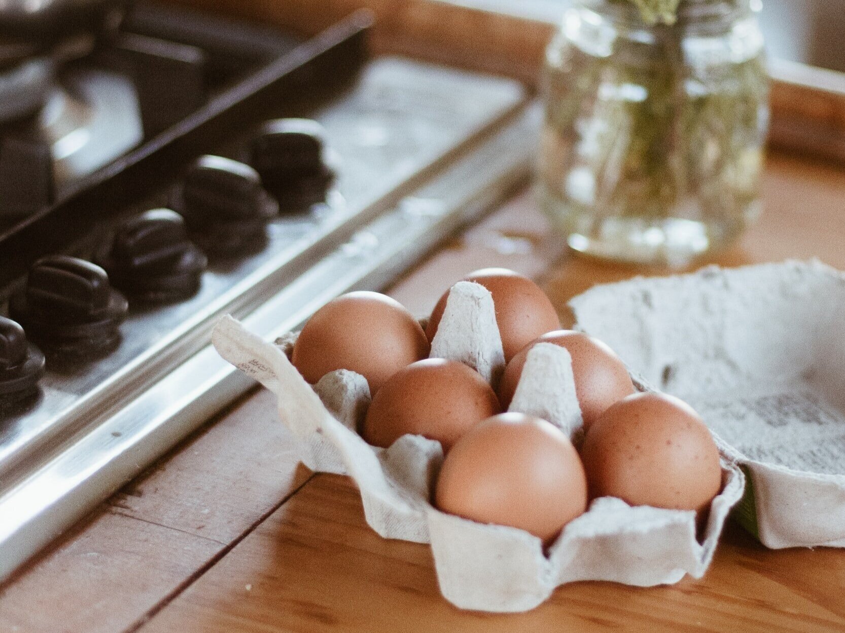 Fresh brown eggs in a cardboard carton on a wooden kitchen countertop, with a gas stove and a jar of herbs in the background. Perfect for cooking and baking.