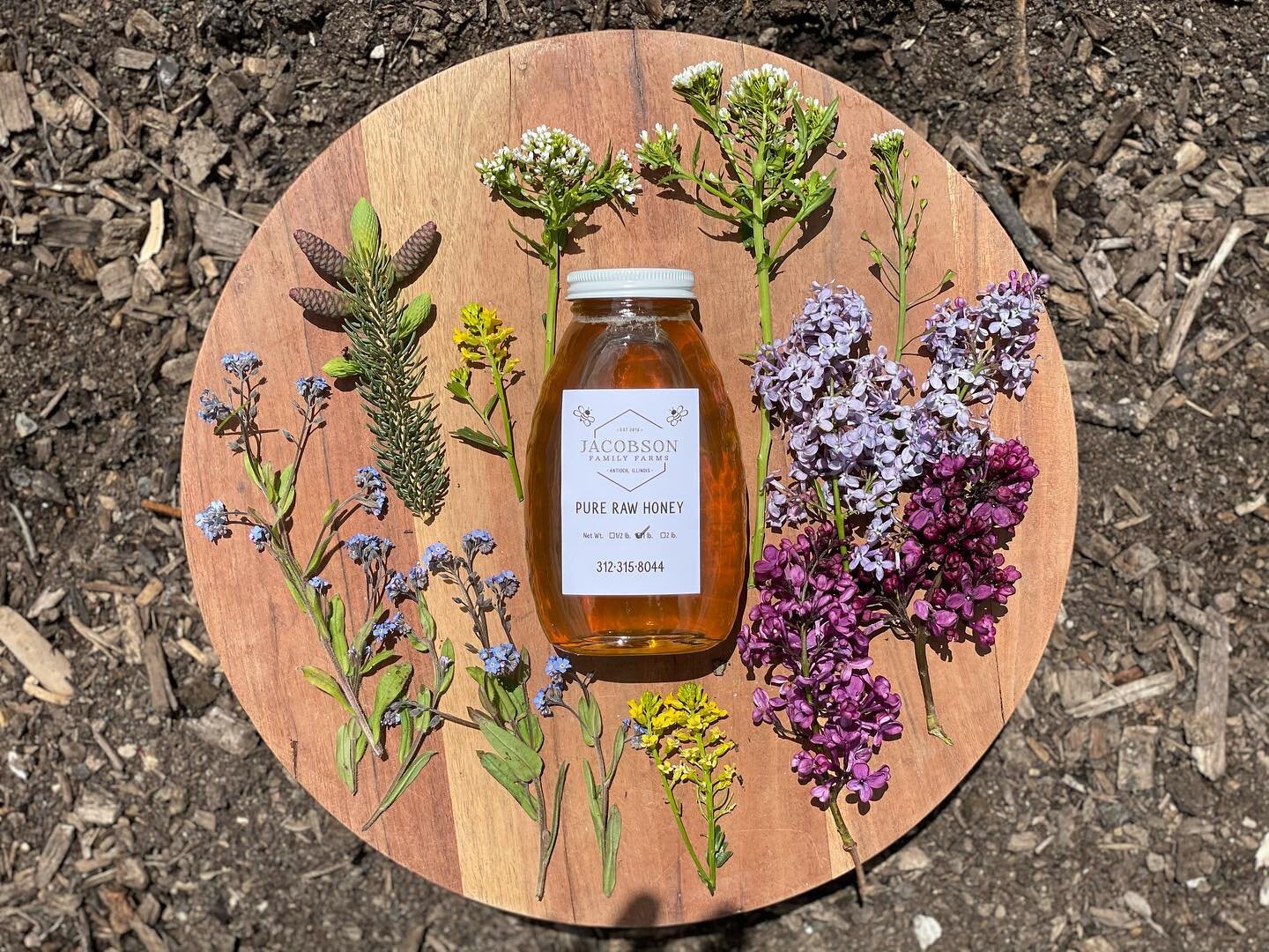 image-asset - 2026-07-23T084226.612 A bottle of Jacobson Pure Raw Honey surrounded by colorful wildflowers and greenery on a wooden platter, set against a natural soil background, showcasing a rustic and organic aesthetic.