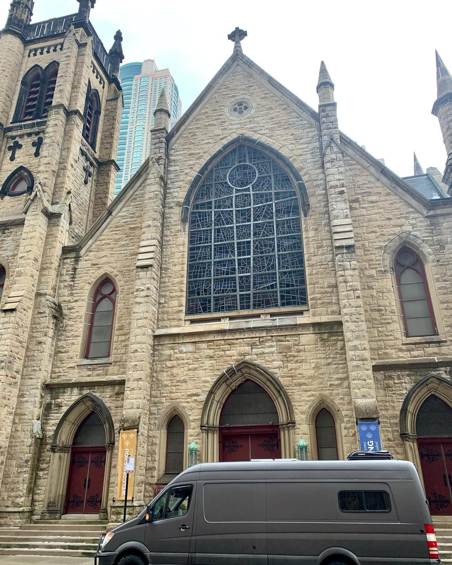 image-asset - 2026-07-23T084228.431 Historic stone church facade featuring intricate stained glass windows, tall spires, and a modern van parked in front, with a backdrop of skyscrapers in an urban setting.