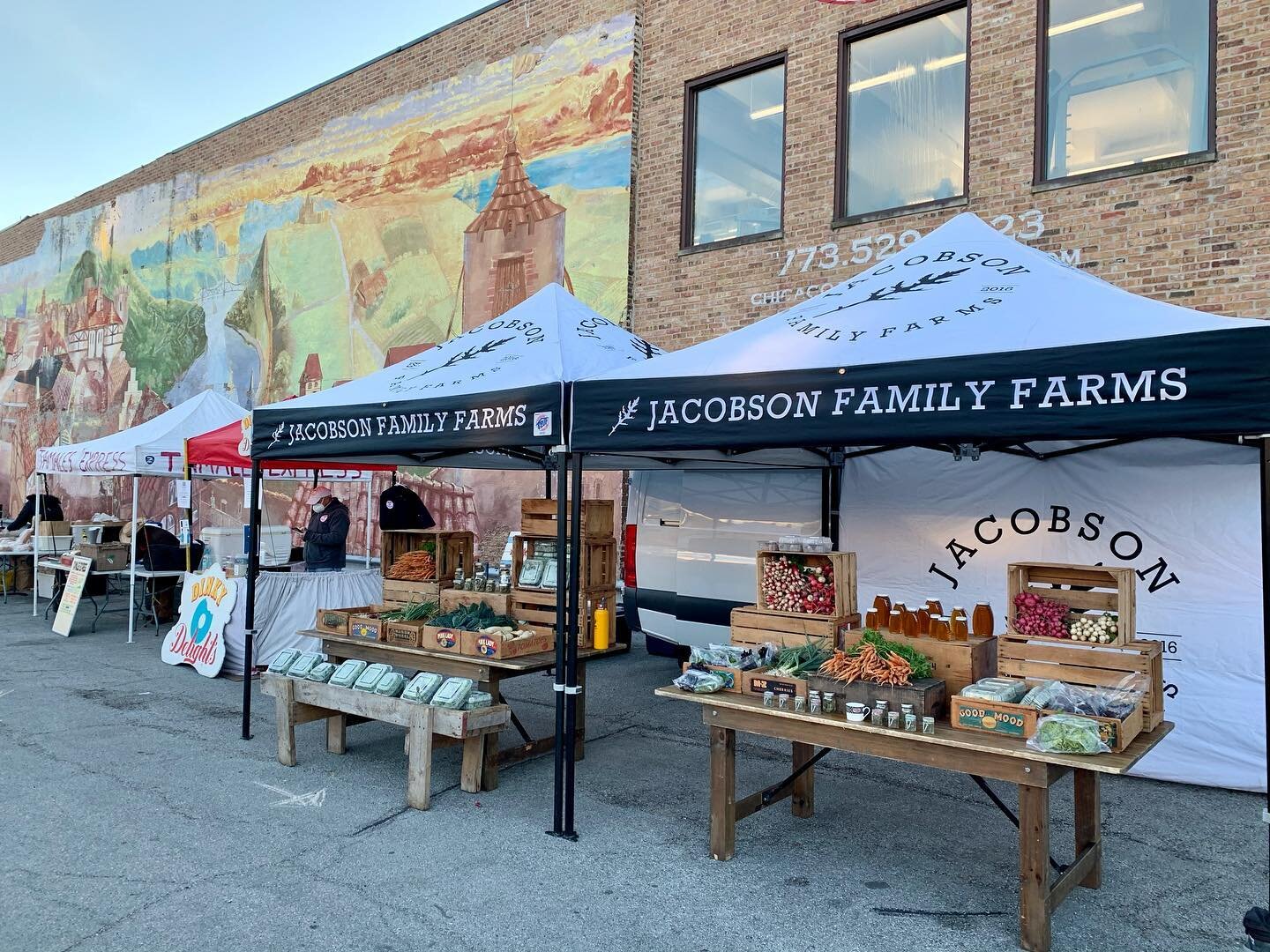 image-asset - 2026-07-23T084241.040 Jacobson Family Farms market stall featuring fresh produce, jars of homemade goods, and vibrant vegetables. The backdrop showcases a colorful mural, enhancing the outdoor market atmosphere. Perfect for local food enthusiasts and community events.