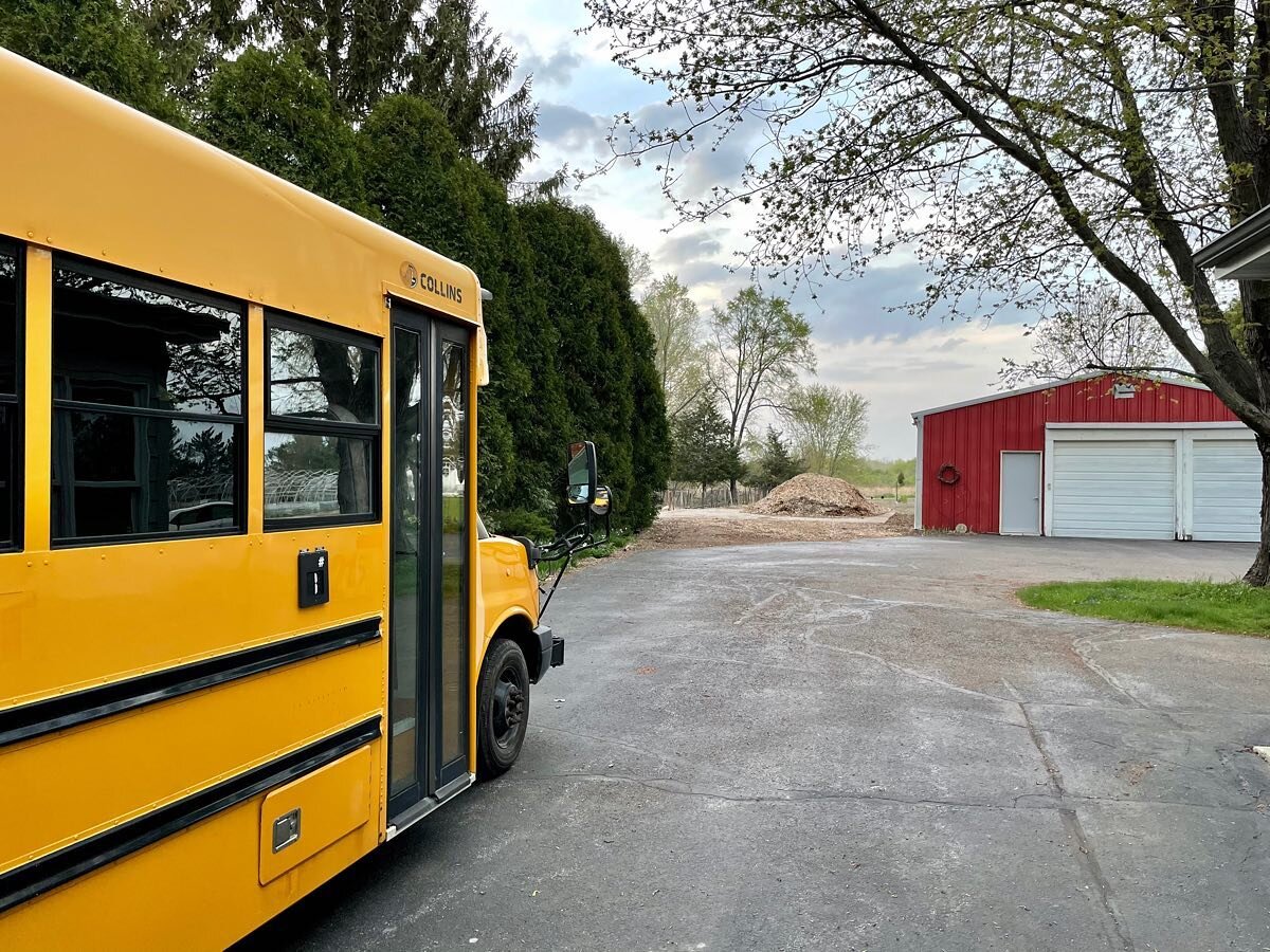image-asset - 2026-07-23T084244.597 Yellow school bus parked on a residential driveway with a red barn in the background and a pile of wood chips nearby, surrounded by greenery and cloudy skies.